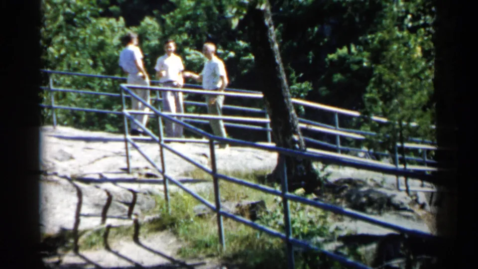 The 1961 Minnesota home-movie frame shows three men in pale clothing talking beside a railing, with trees and exposed stone around them.