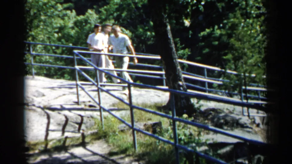 At the end of the 1961 Minnesota clip, three adult men remain near the blue railing on sunlit rock surrounded by dense green trees.