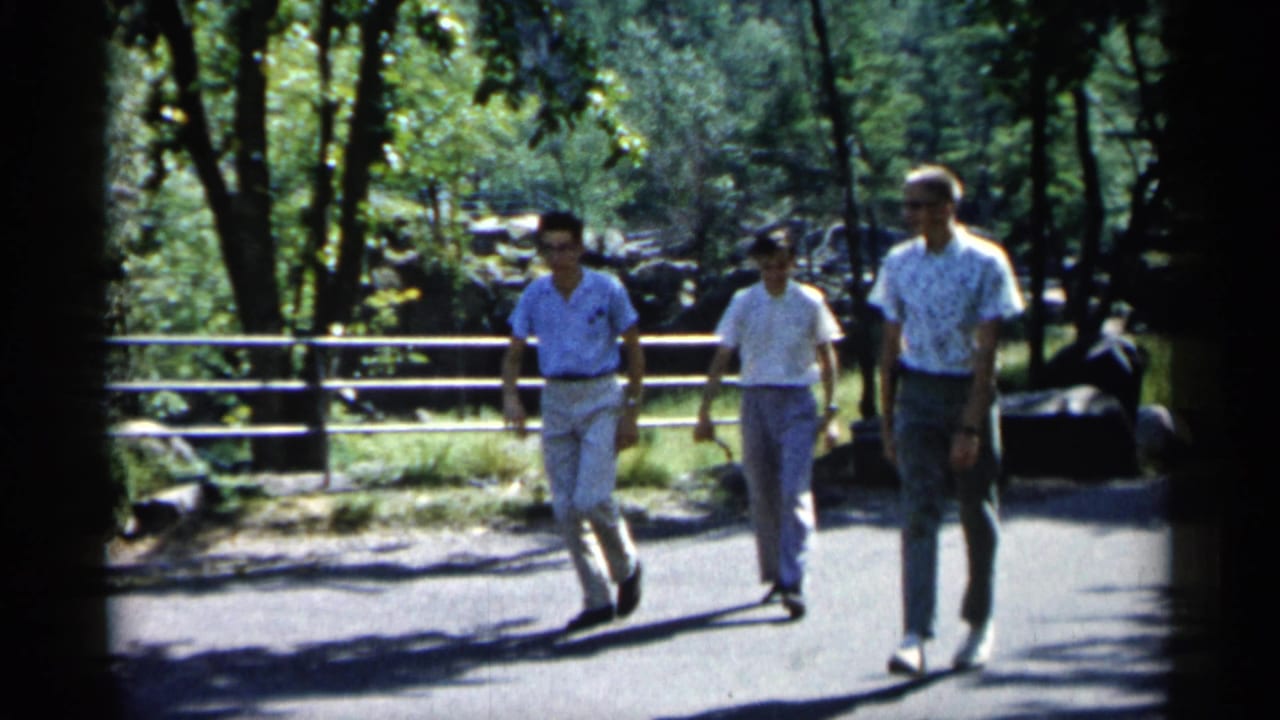 Three men continue walking on a bright park path beside railings.