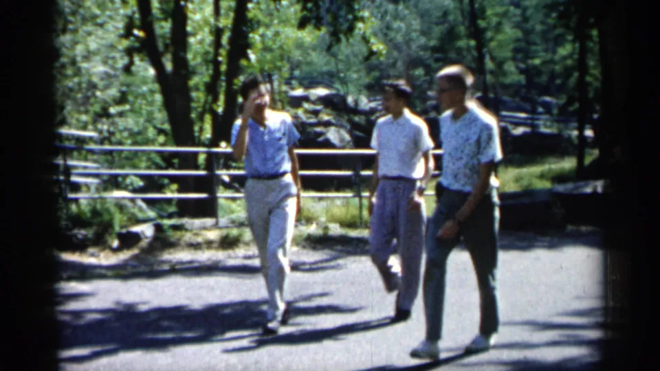 Three men in early-1960s casual attire pass through the frame on a tree-lined Minnesota park path.