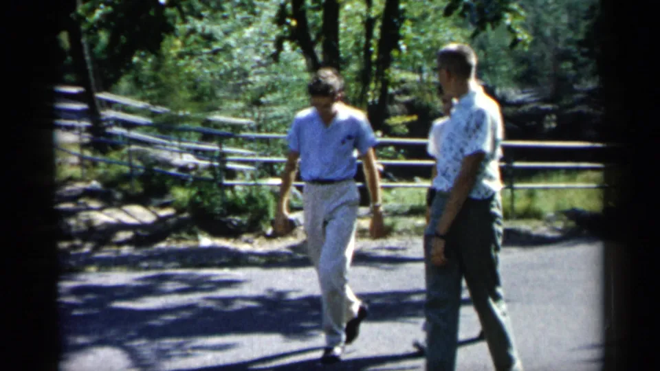Two of the men are visible near the end of the shot, walking through a shaded 1961 Minnesota park scene.