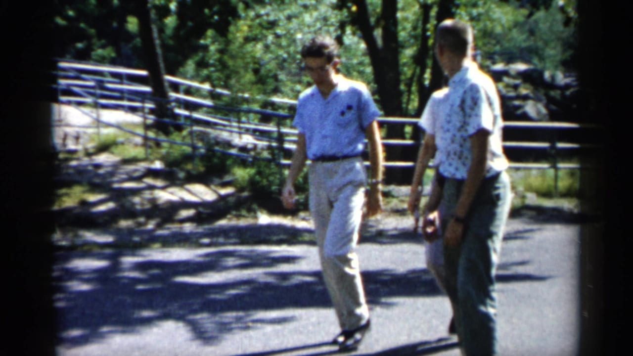 Two men walk on a paved path in a leafy park with railings behind them.