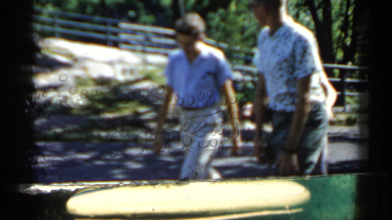 Two men pass near a railing in a park, partly obscured by soft focus and film gate edges.