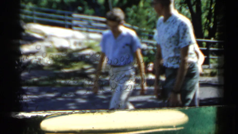Two men in patterned and light shirts walk past park railings in Minnesota, captured in soft, handheld 8mm framing.