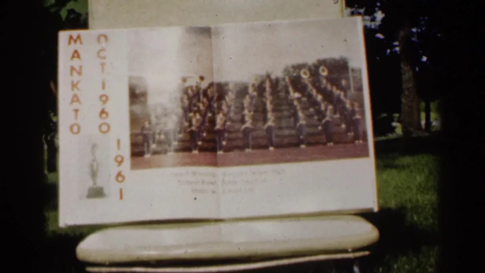 The Mankato program remains framed outdoors, showing a marching band photograph, vertical date text, and shaded park background.