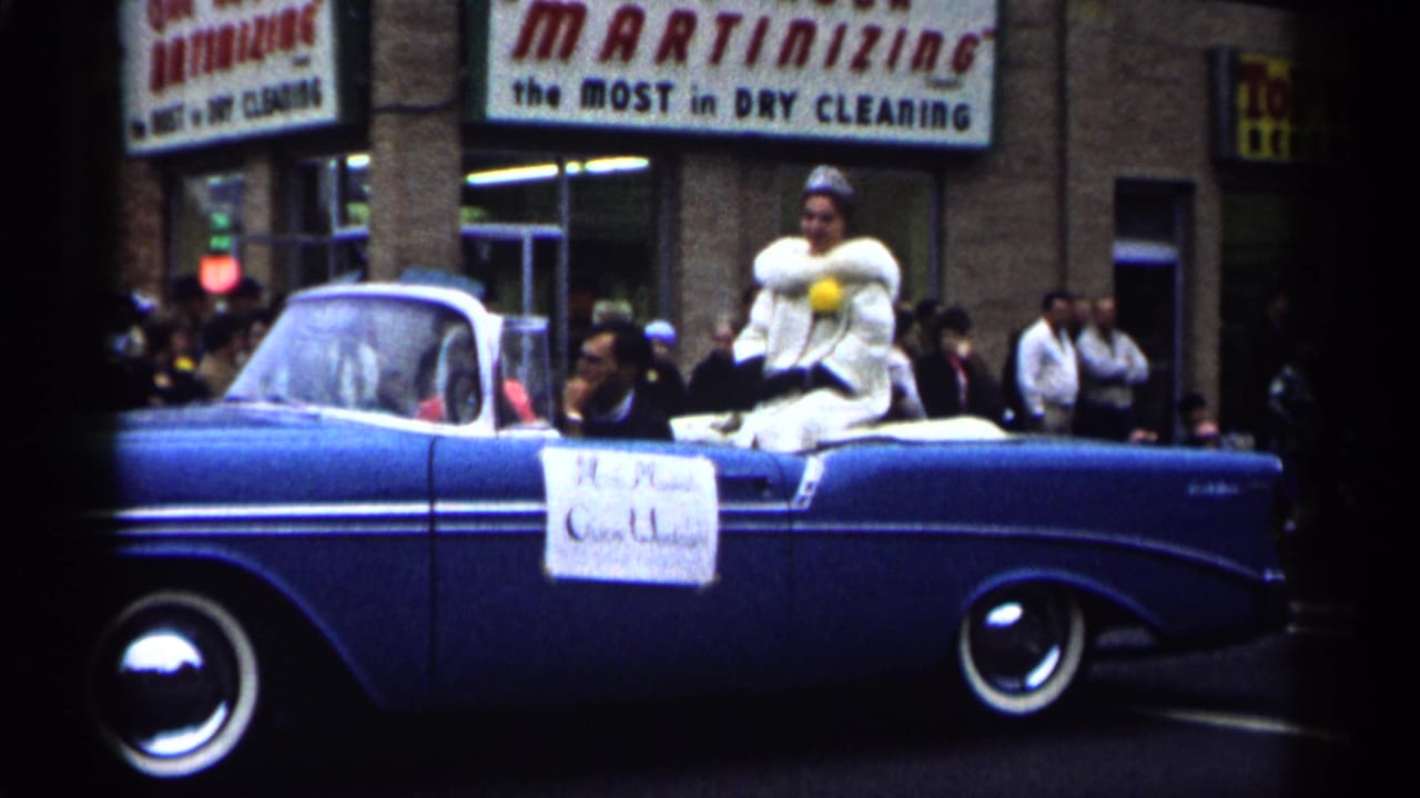 1961 Minnesota parade view of a blue convertible carrying a woman in a white fur-trimmed outfit past downtown storefronts.