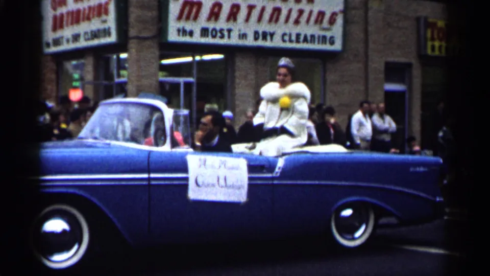 1961 Minnesota parade view of a blue convertible carrying a woman in a white fur-trimmed outfit past downtown storefronts.
