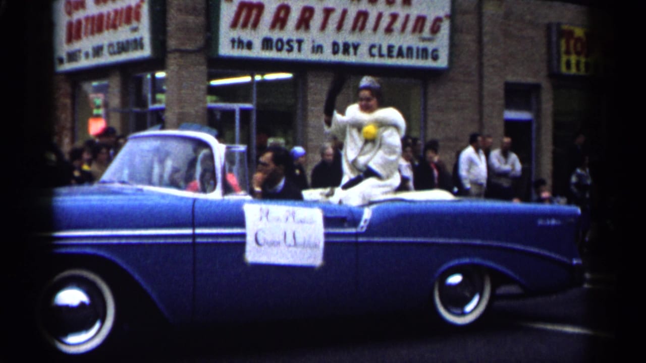 Woman waves from the rear of a blue convertible as the parade car passes storefront spectators.