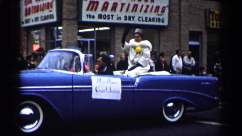 A woman in white parade attire waves from a blue convertible on a Minnesota street, with storefront signs and onlookers behind her.