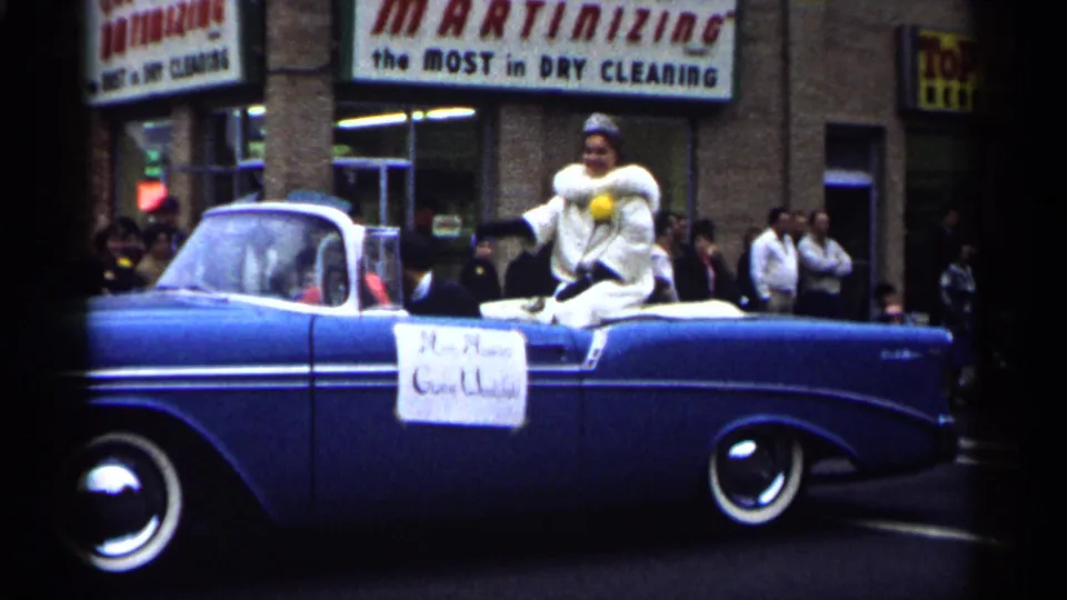 The blue parade convertible continues past a storefront, with the woman seated at the rear and a sign attached to the car door.
