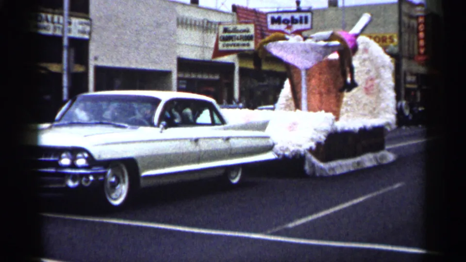 A white 1960s car pulls a parade float resembling a giant cocktail glass, with fluffy trim and a pink umbrella visible on the street.