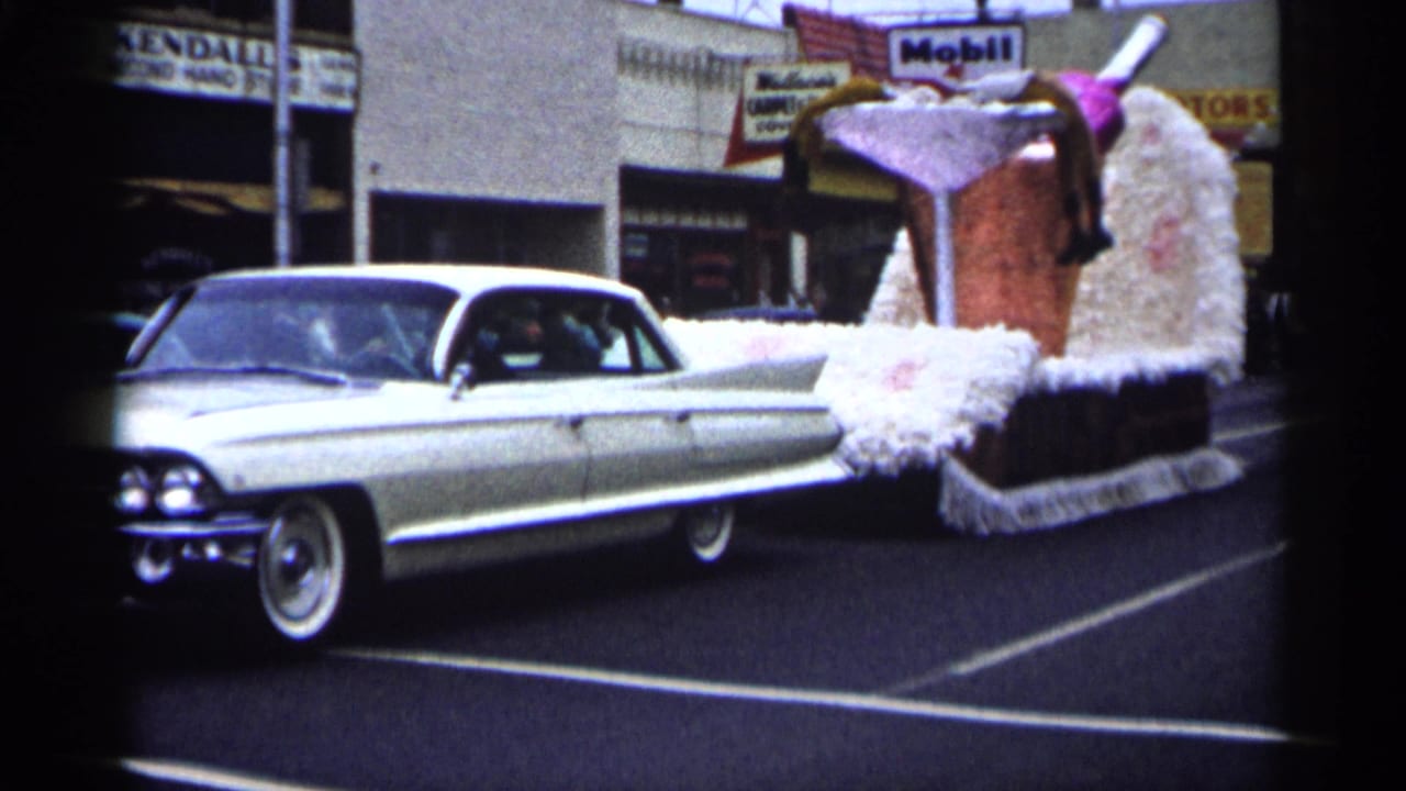 White Cadillac pulling a giant cocktail-themed parade float past storefronts.