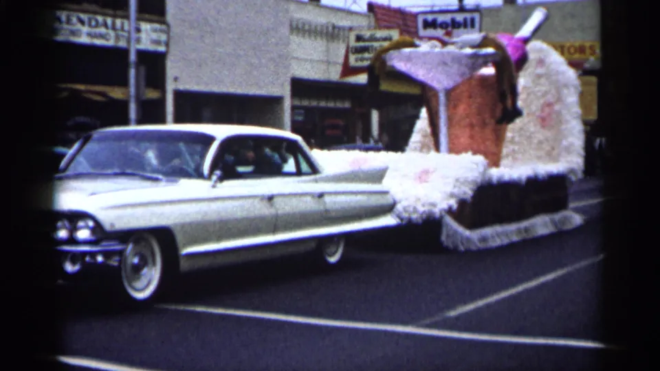 1961 Minnesota home-movie frame showing a white Cadillac pulling a large martini-glass parade float along a city street.