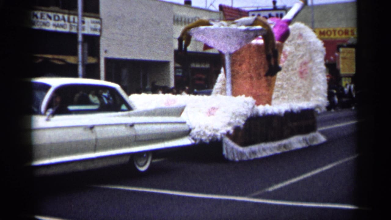 Cadillac and cocktail float moving past Mobil and second-hand store signs.