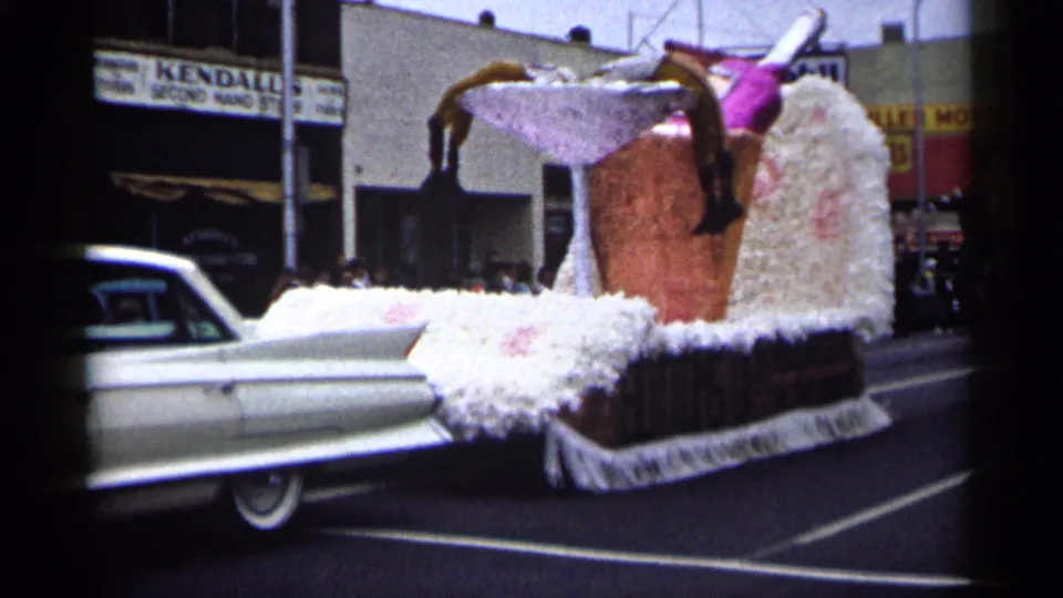 A close curbside view centers the tall cocktail float, with the white Cadillac partly leaving the frame on a 1961 Minnesota street.