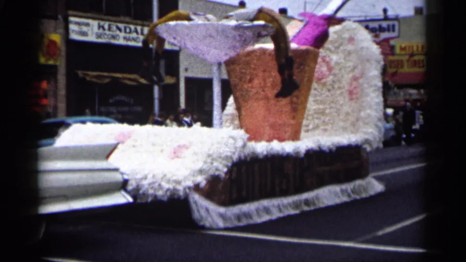 The parade float, trimmed in white and topped with oversized drink props, passes storefronts and spectators in Minnesota in 1961.