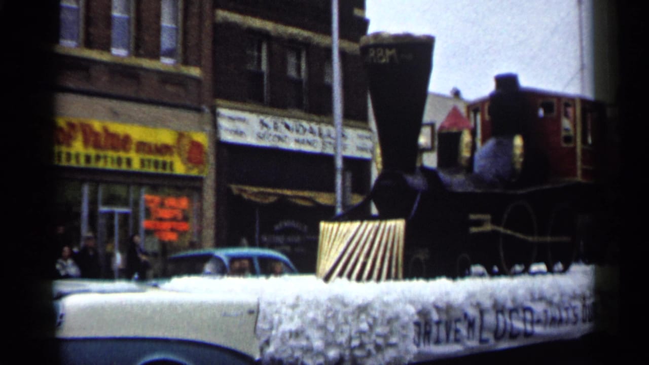 1961 Minnesota parade frame showing a black-and-red steam train float beside storefronts, cars, and visible Mobil and Top Value signage.