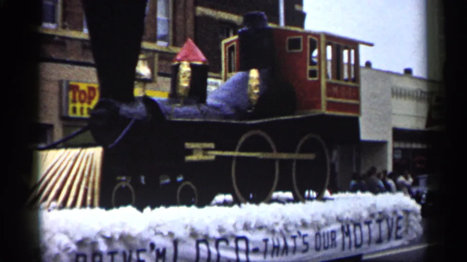 1961 Minnesota parade frame showing the side of a black-and-red train float with white fringe and storefronts in the background.