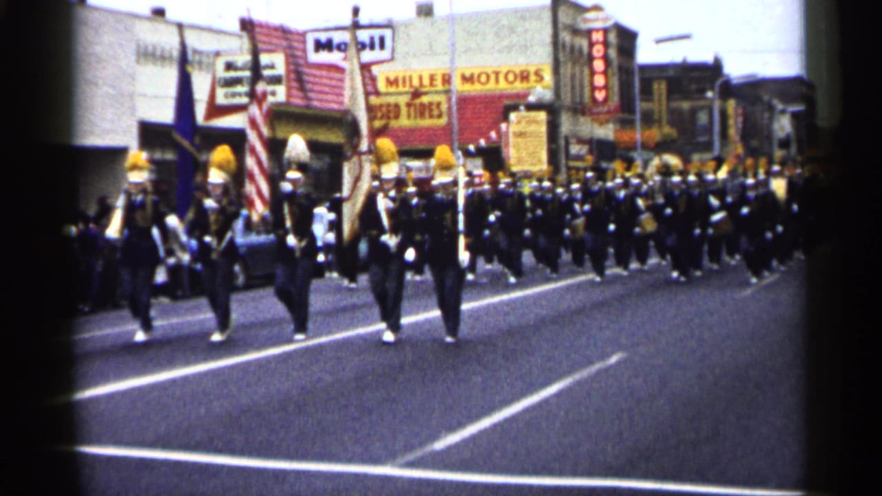 Marching band approaches on a town street with flags and storefront signs behind them.