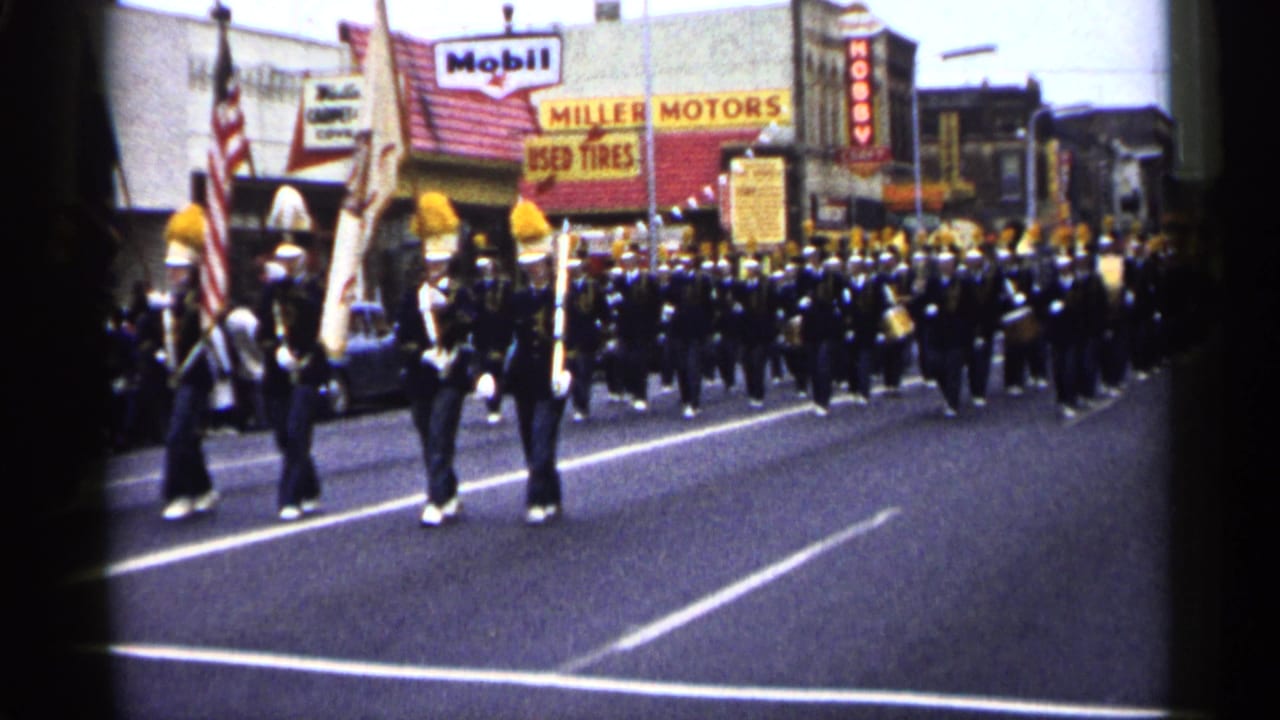 Uniformed marching band fills the street as flags pass near the camera.
