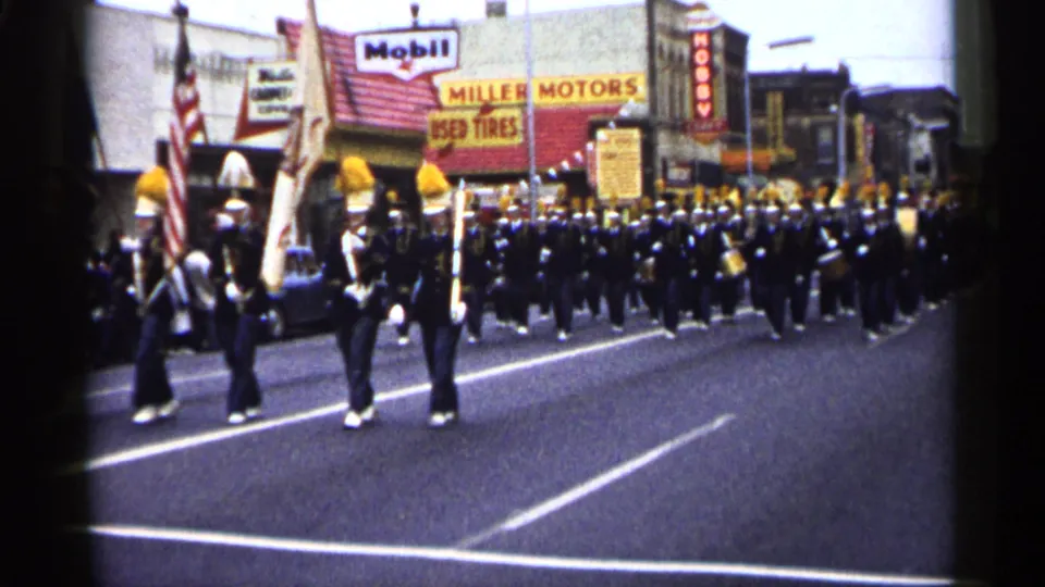 Large black-and-yellow uniformed marching band continues along a Minnesota street in 1961, with storefront and gas-station signs visible.