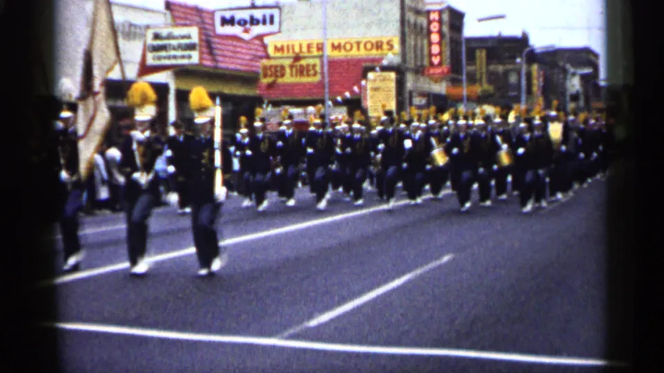 Curbside 8mm view of uniformed band members and a flag bearer passing storefronts during a 1961 Minnesota parade.