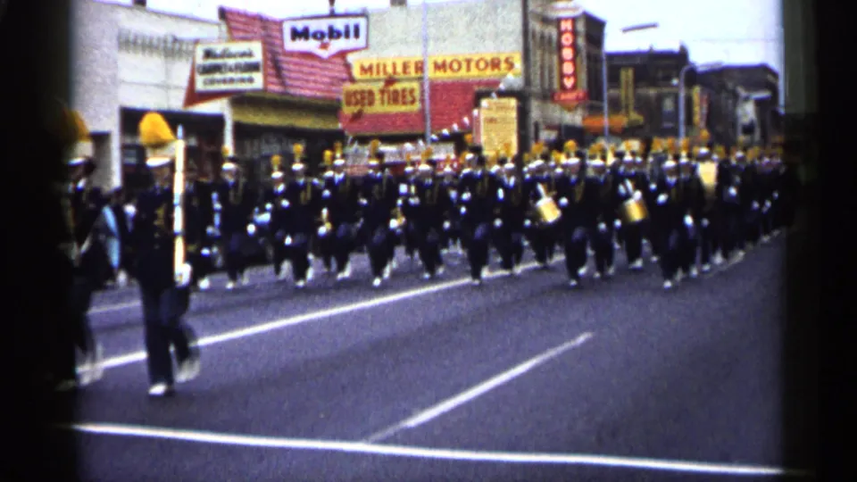 Rows of marching musicians in black-and-yellow uniforms pass the camera on a Minnesota street, with Mobil and Miller Motors signs behind them.