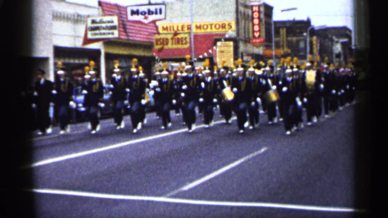 Uniformed marching band advances along the street with drums and brass near storefronts.