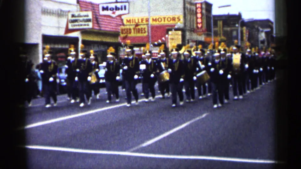 1961 Minnesota home-movie frame of a marching band spread across a town street, with Miller Motors and used tire signage visible.