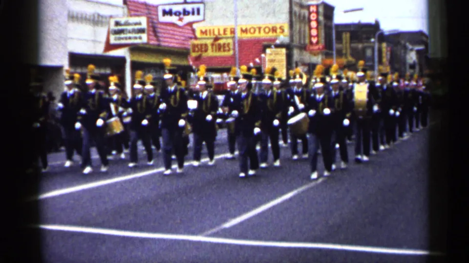 1961 Minnesota parade frame showing uniformed musicians with drums and brass on a street bordered by Mobil and Miller Motors signage.