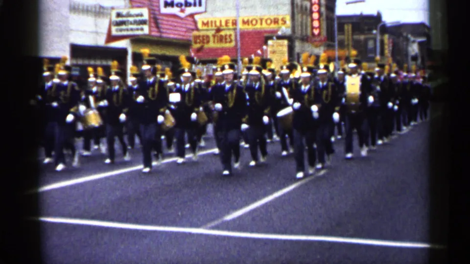 1961 Minnesota street parade footage showing a marching band in dark uniforms with yellow plumes approaching the camera.