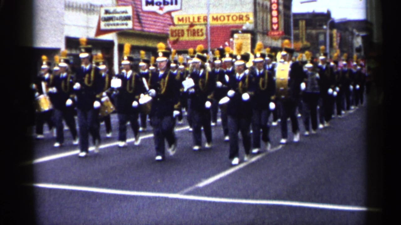 Band members march forward in formation with drums and brass instruments on a paved street.