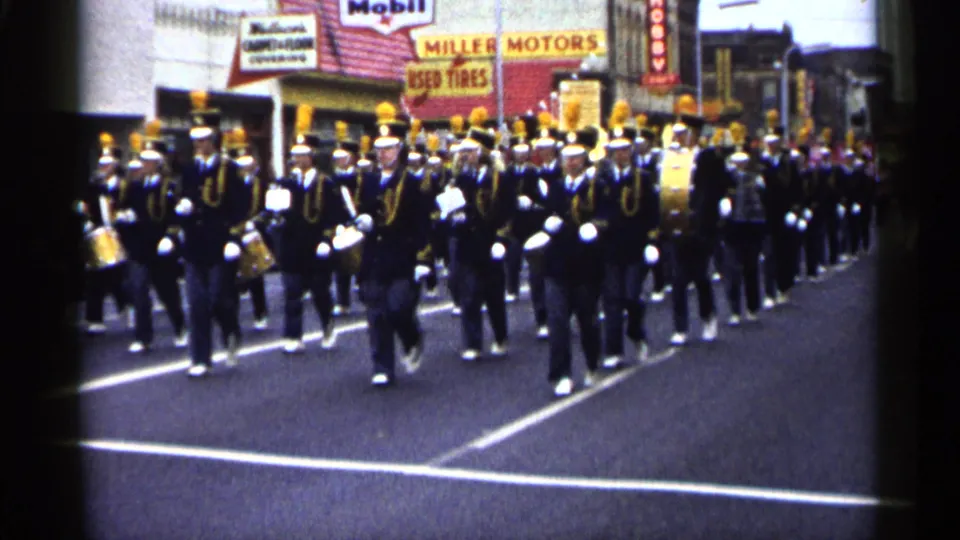 Uniformed musicians in a Minnesota parade move closer to the camera, with drums, brass instruments, and storefront signs visible.