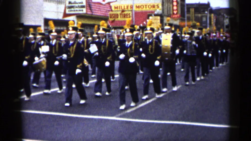 Closer 8mm frame of the 1961 Minnesota marching band, with gold-trimmed uniforms, white gloves, drums, and street signs behind them.