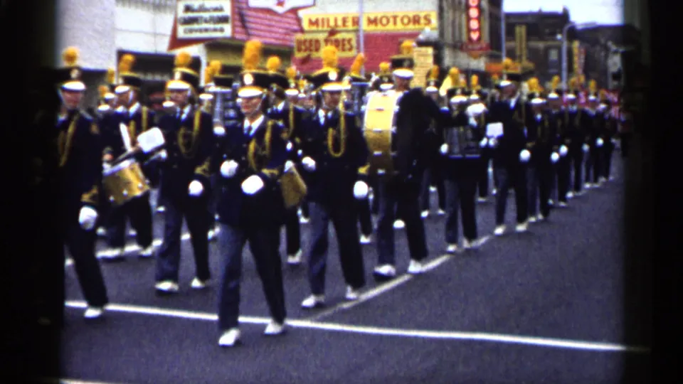The marching band passes near the camera in 1961 Minnesota footage, with plumed hats, drums, and visible commercial signs in the background.