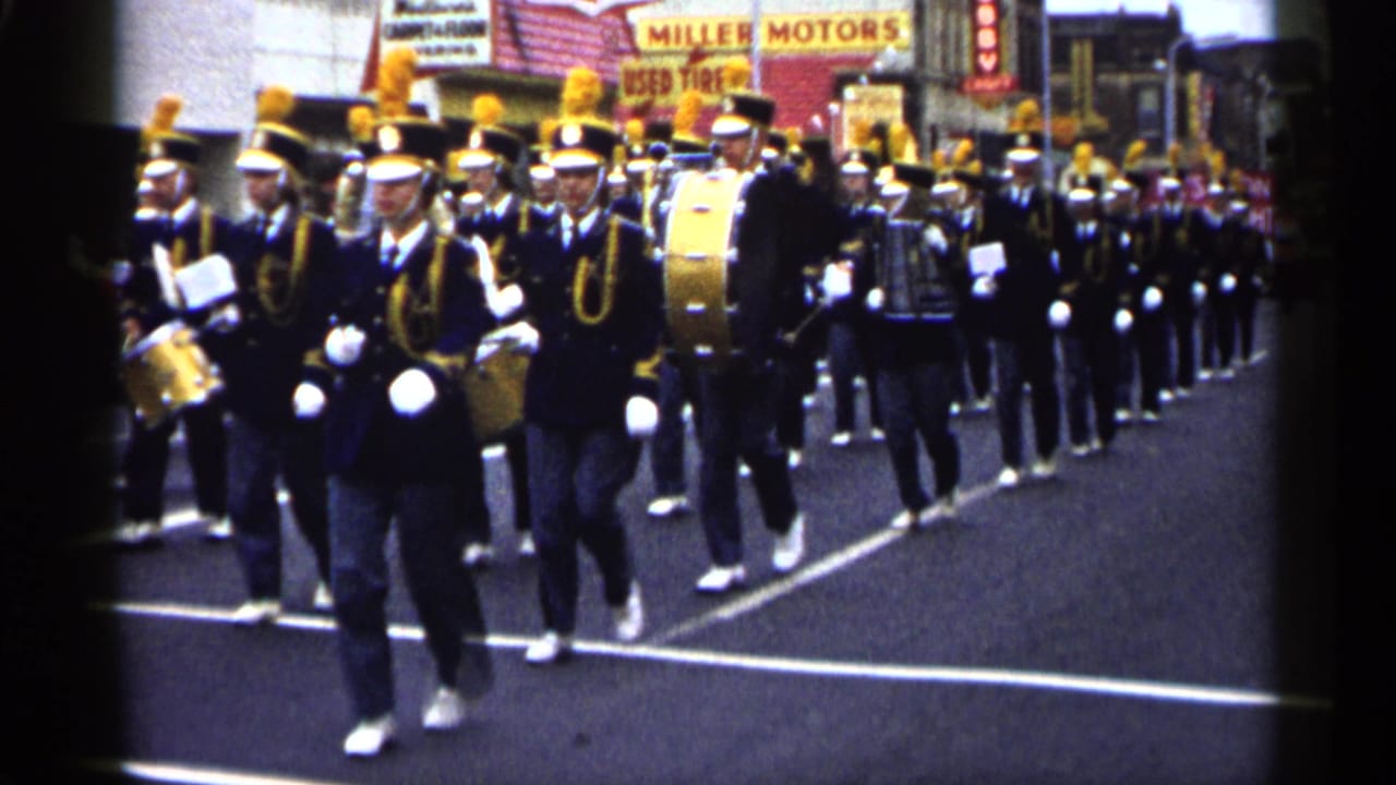 Marching band in dark uniforms advances down a city street past storefront signs.