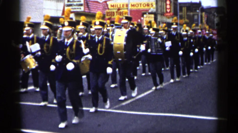 1961 Minnesota home-movie frame of a marching band in plumed hats moving toward the camera on a commercial street.
