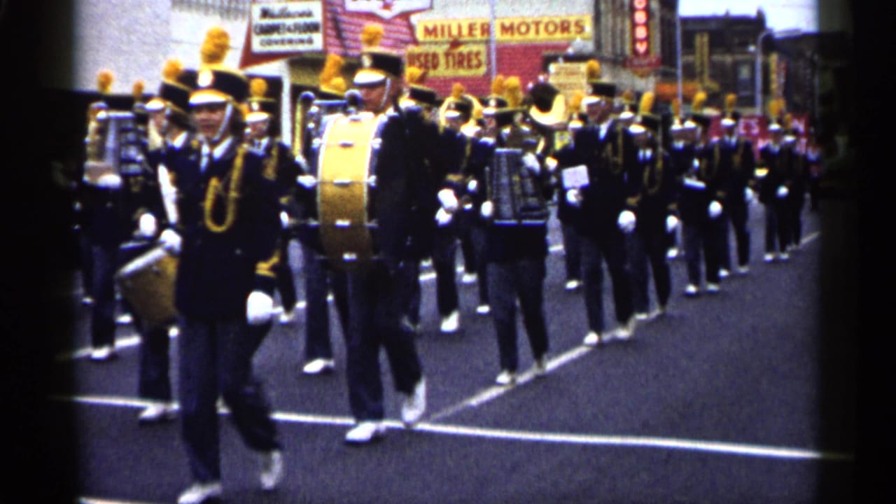 Marching musicians in dark uniforms and tall plumed hats pass the camera during a 1961 Minnesota street parade.