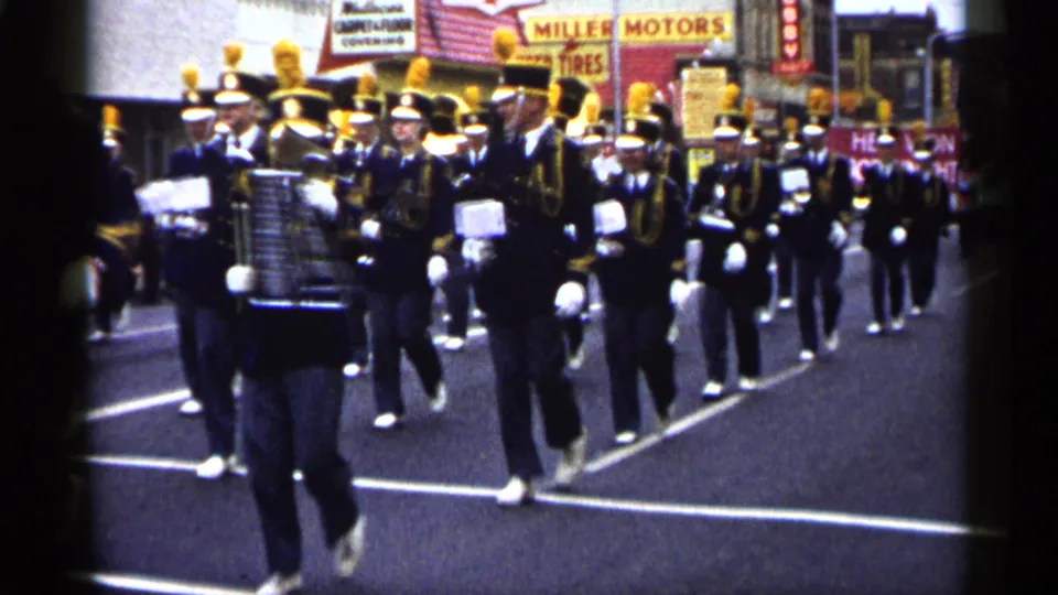 1961 Minnesota parade frame with percussion players and uniformed band members marching past storefront signs.