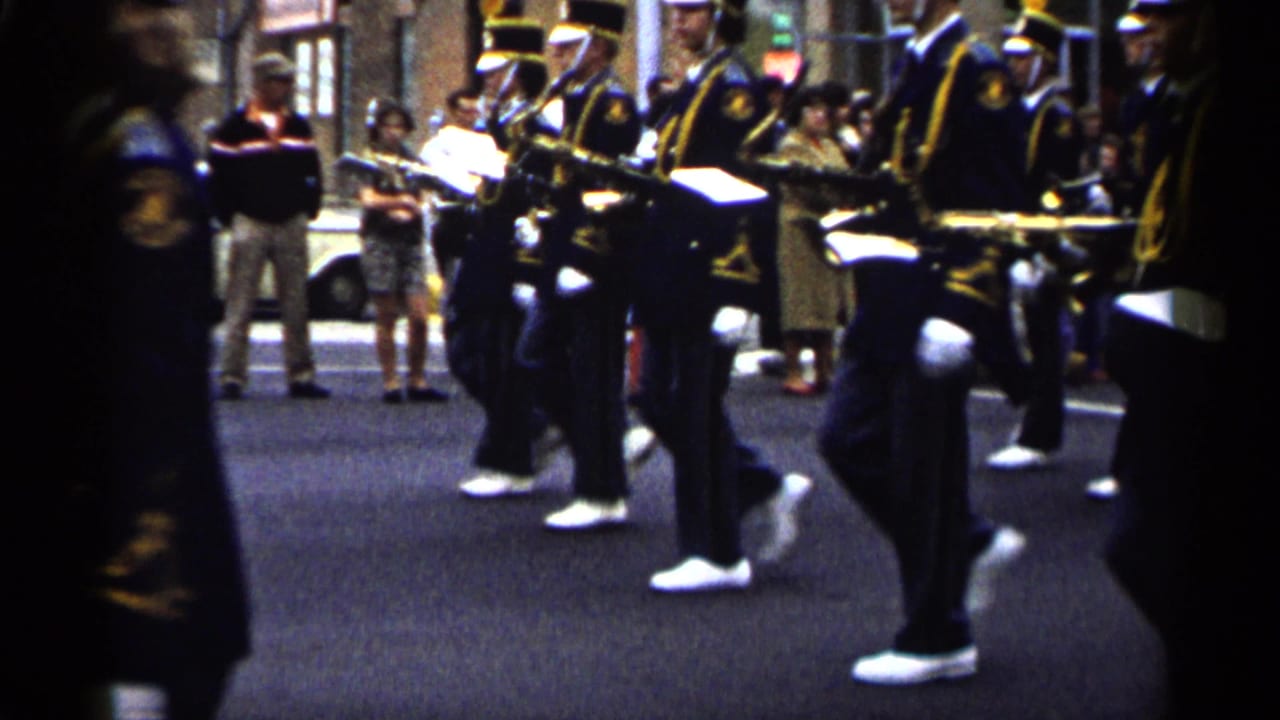 1961 Minnesota 8mm frame showing a marching band in dark uniforms with gold trim passing storefronts and sidewalk spectators.