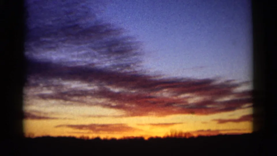 1961 Minnesota 8mm frame with a bright orange-yellow sunset glow below broad blue clouds and a dark rural horizon line.