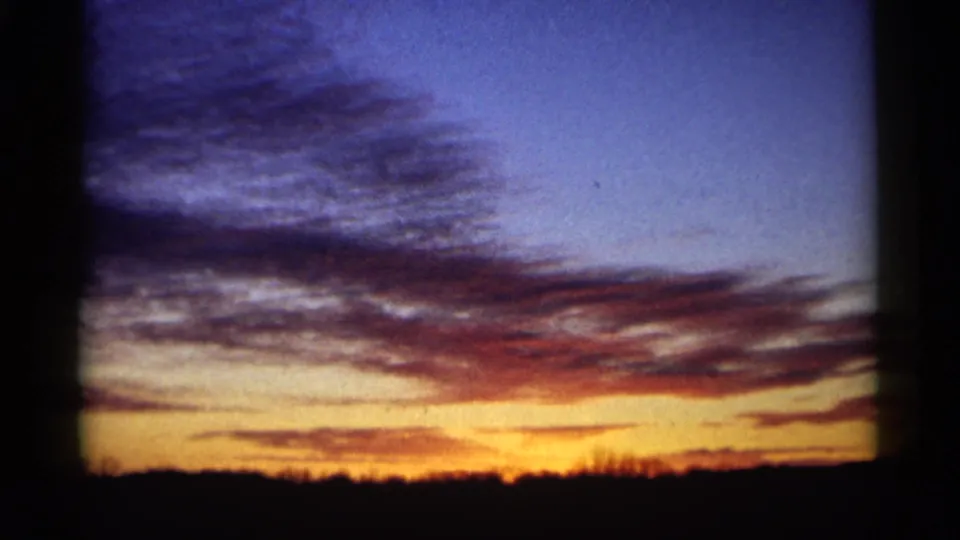 1961 Minnesota 8mm sunset frame showing blue dusk clouds, orange light near the horizon, and a silhouetted treeline below.