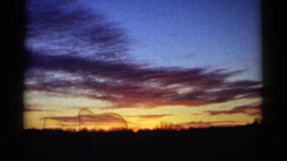 1961 Minnesota 8mm frame of a sunset sky with reddish cloud bands and a dark horizon, including a faint translucent film or lens mark near the lower sky.