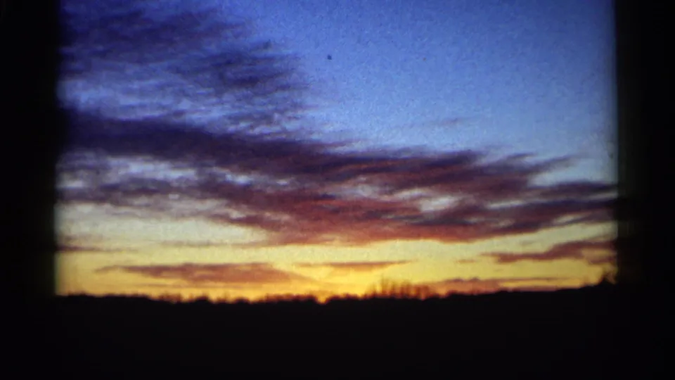 1961 Minnesota 8mm frame with layered blue and purple clouds over a glowing sunset and low dark horizon.