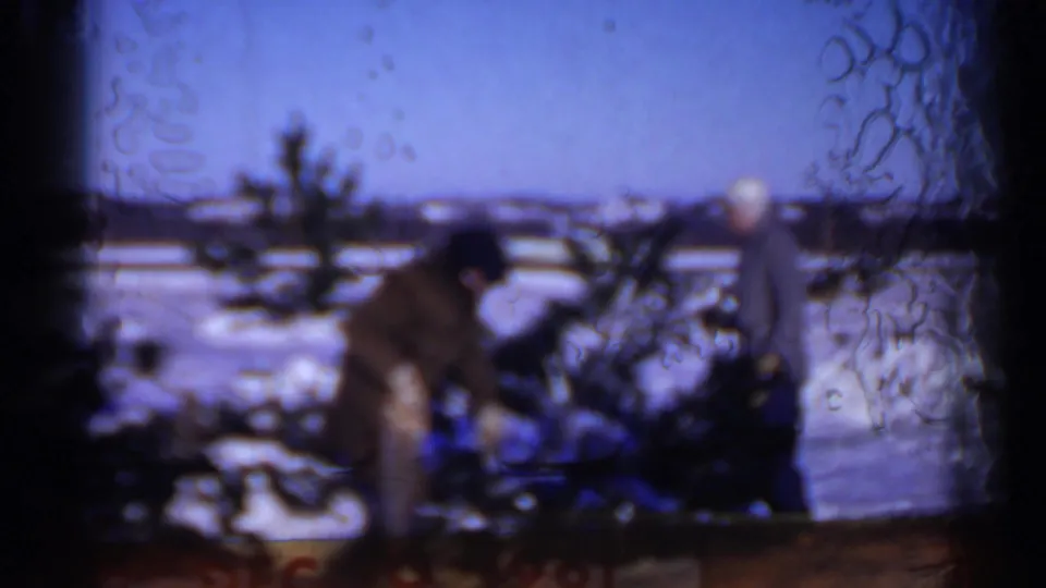 Soft 8mm frame from Minnesota in 1961 showing a man bending near a pine tree in snow, with another person partly visible beyond.