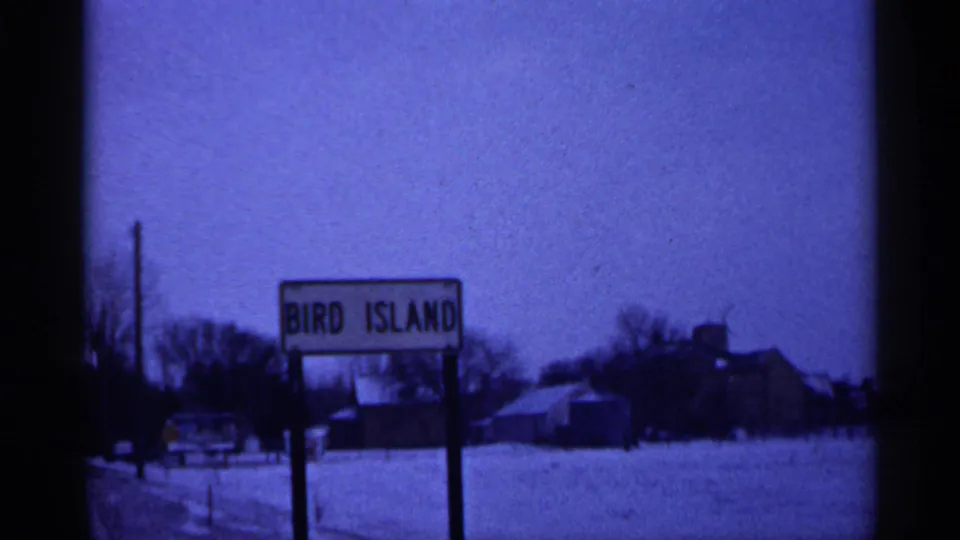 Static 1961 winter view of the Bird Island sign in Minnesota, with snow on the ground, bare trees, and rural buildings beyond.