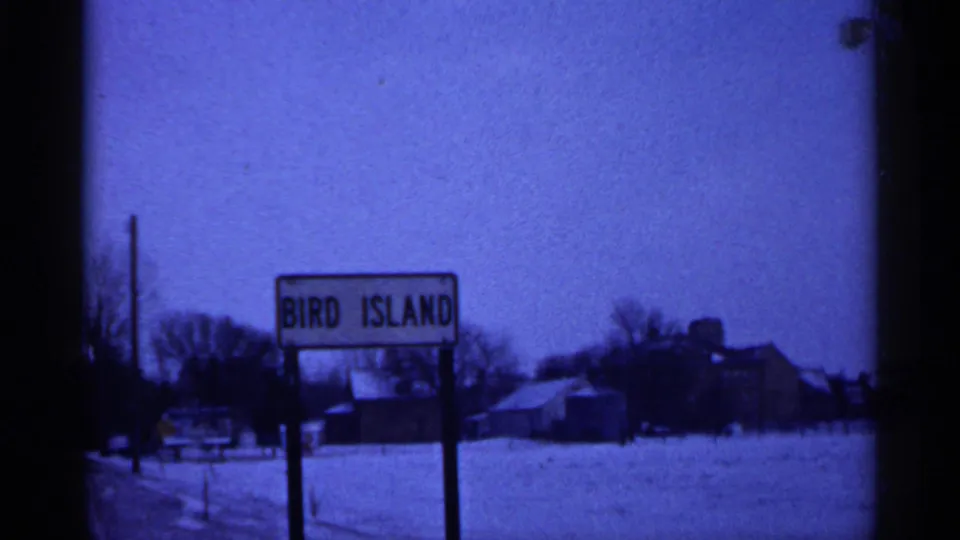 Blue-tinted 8mm frame of the Bird Island sign in a quiet Minnesota winter landscape with snow, bare trees, and farm-like buildings.