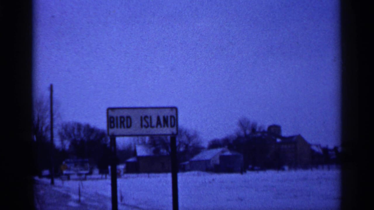1961 Minnesota 8mm frame showing a Bird Island roadside sign in a snowy rural setting with distant buildings and bare trees.