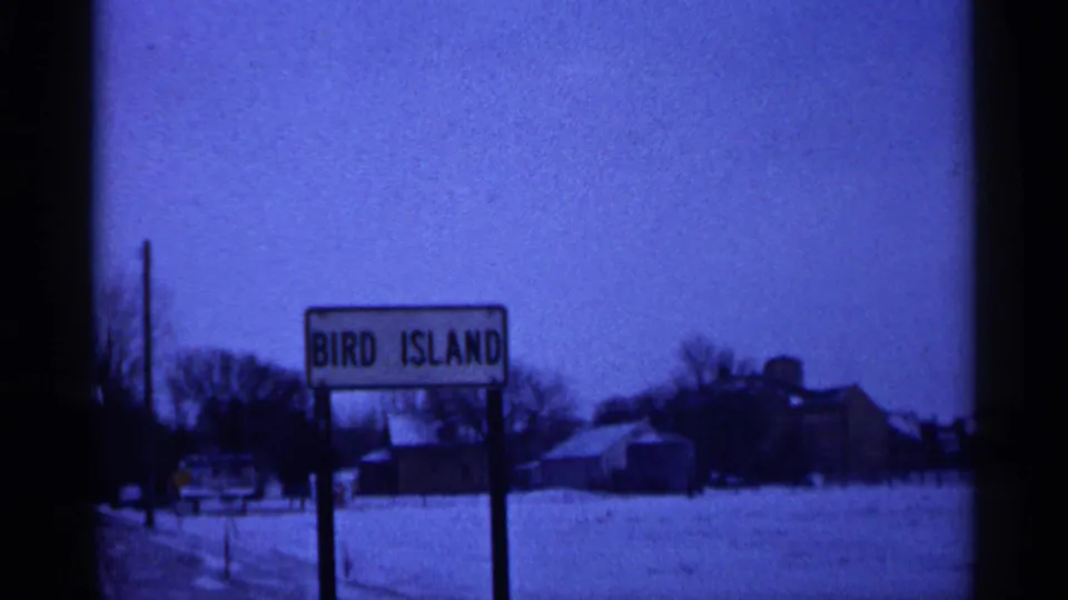 1961 Minnesota 8mm frame showing a Bird Island roadside sign in a snowy rural setting with distant buildings and bare trees.