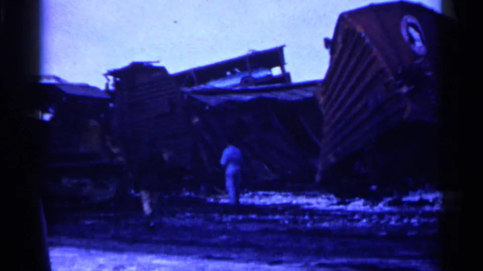 1961 Minnesota 8mm frame of overturned rail cars and wreckage in a winter field, with an adult bystander near the scene.